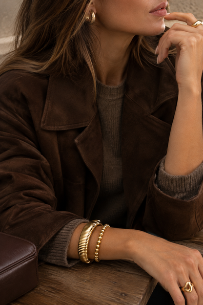 Audace Wide Cuff in gold stacked with a gold bead bracelet, worn with a brown suede jacket