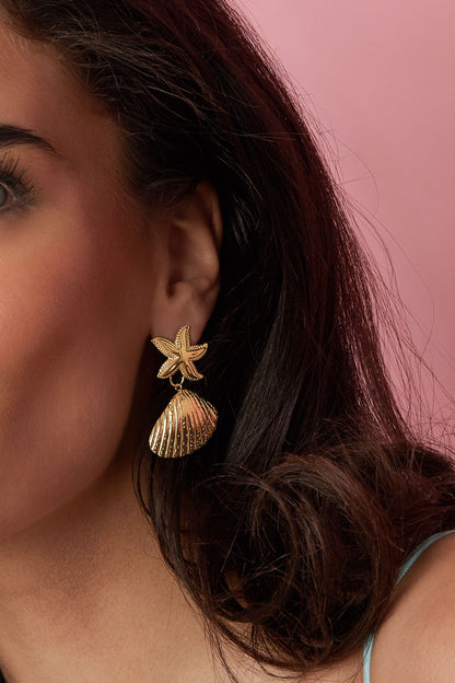 Close-up of a woman wearing gold starfish and shell earrings against a pink background